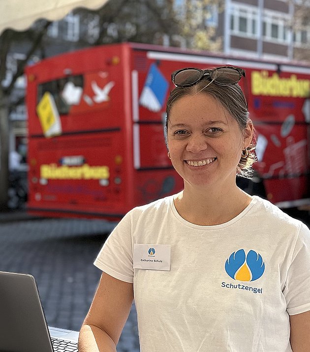 Zwei Personen mit weißen "Schutzengel"-T-Shirts stehen an einem Stand im Freien unter einem Zelt. Der Stand ist mit Broschüren, Testkits und einem Laptop ausgestattet. Im Hintergrund sind ein "Schutzengel"-Transparent und ein roter Lastwagen zu sehen.