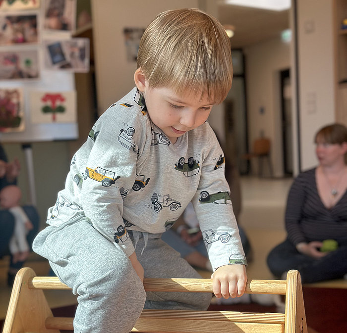 Ein kleines Kind in einem grauen Schlafanzug mit Autoaufdrucken klettert in einem Haus auf eine Holzleiter. Das Sonnenlicht scheint auf das Kind, während im Hintergrund Erwachsene auf einem roten Teppichboden sitzen.