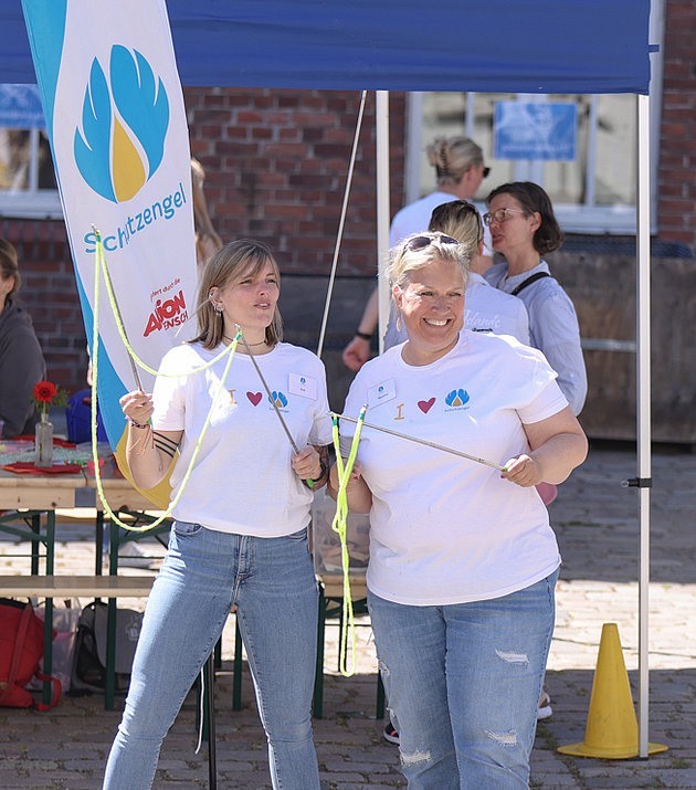 Zwei Frauen in weißen Hemden lächeln und posieren mit einer Flagge bei einer Veranstaltung im Freien, während eine Person ein Foto von ihnen macht. Im Hintergrund sitzen und stehen Menschen unter blauen Zelten an einem sonnigen Tag.