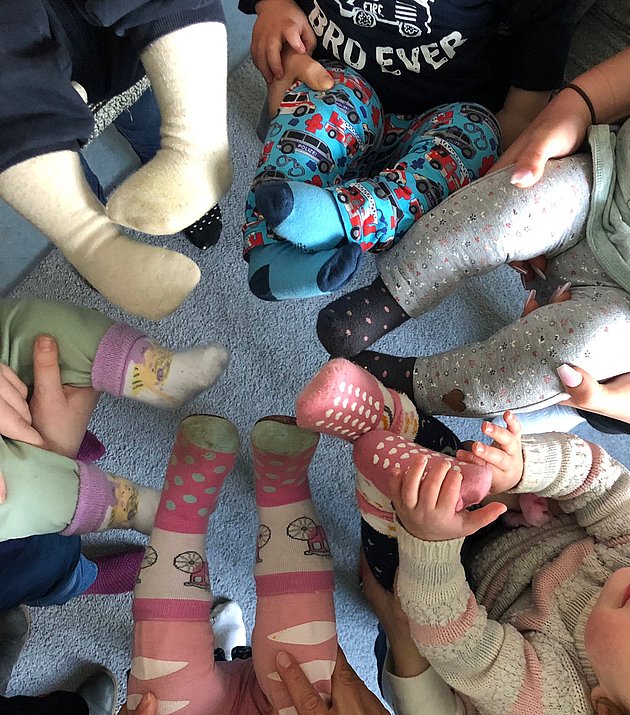 Eine Gruppe kleiner Kinder, die in einem Kreis sitzen, die Füße nach innen gerichtet, mit bunten Socken mit verschiedenen Mustern, auf einem Teppichboden.